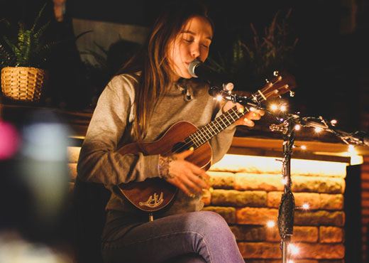 Mulher jovem tocando ukulele e cantando em um cenário noturno com luzes quentes